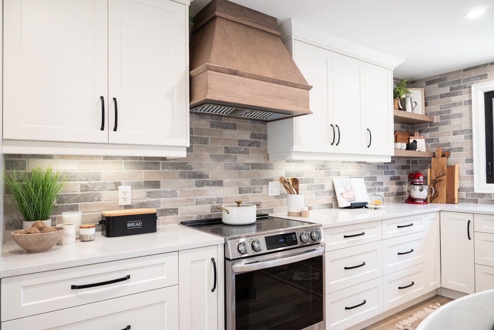 Modern kitchen with white cabinets, gray brick backsplash, stainless steel oven, wooden range hood, open wooden shelves with plants and kitchen items, and natural light from a window.