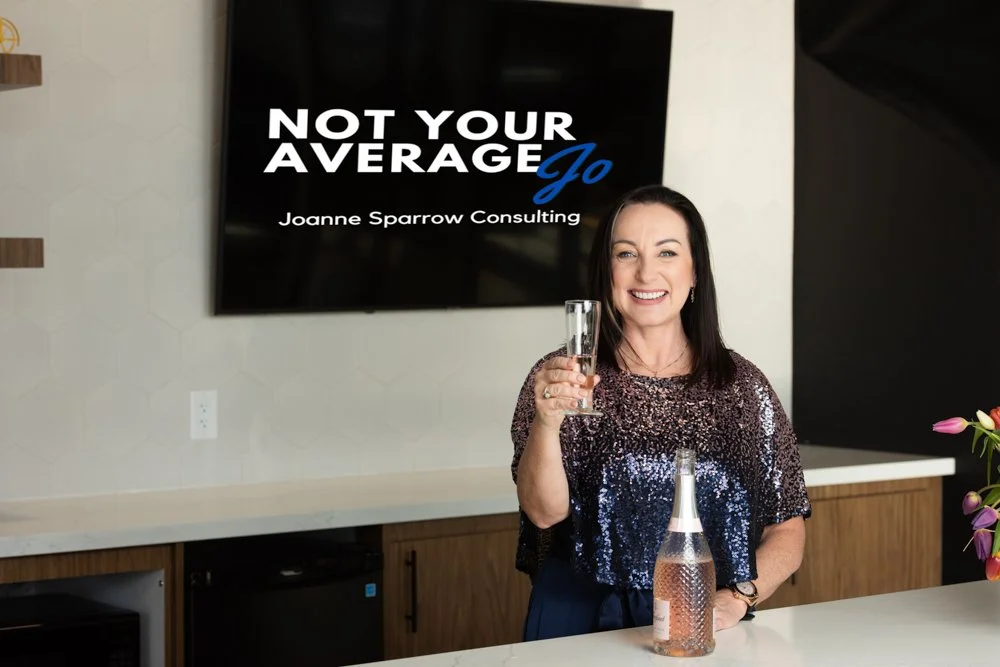 A woman in a sequin top holding a glass of champagne stands behind a counter with a champagne bottle. Behind her, a TV screen displays the text 'Not Your Average Jo' and 'Joanne Sparrow Consulting.'