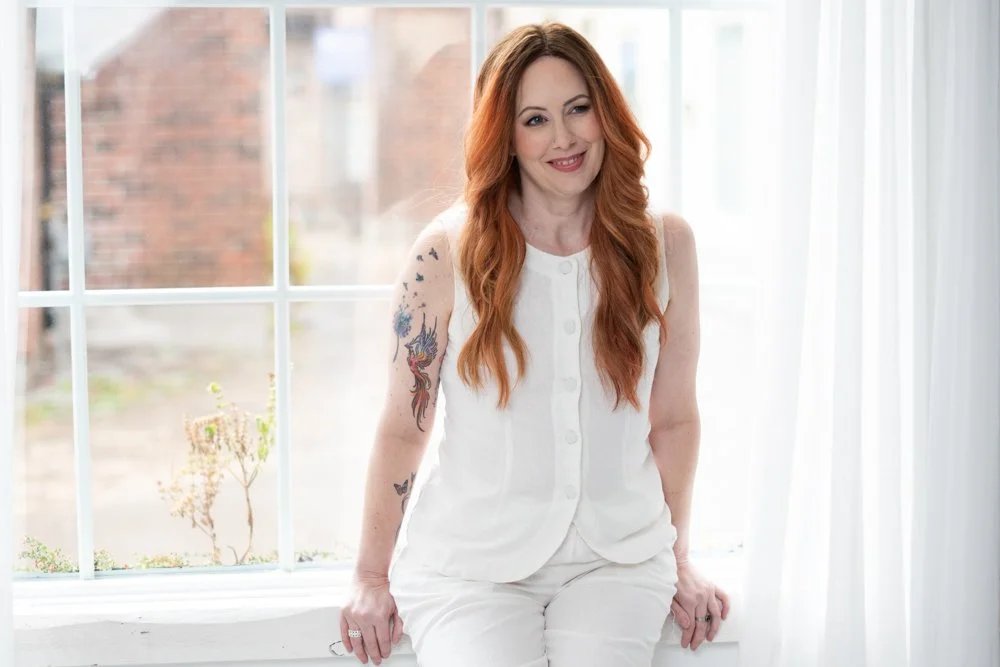 Woman with long red hair and tattoos on her left arm, sitting on a window sill inside a bright room, wearing a white sleeveless top and white pants, smiling.
