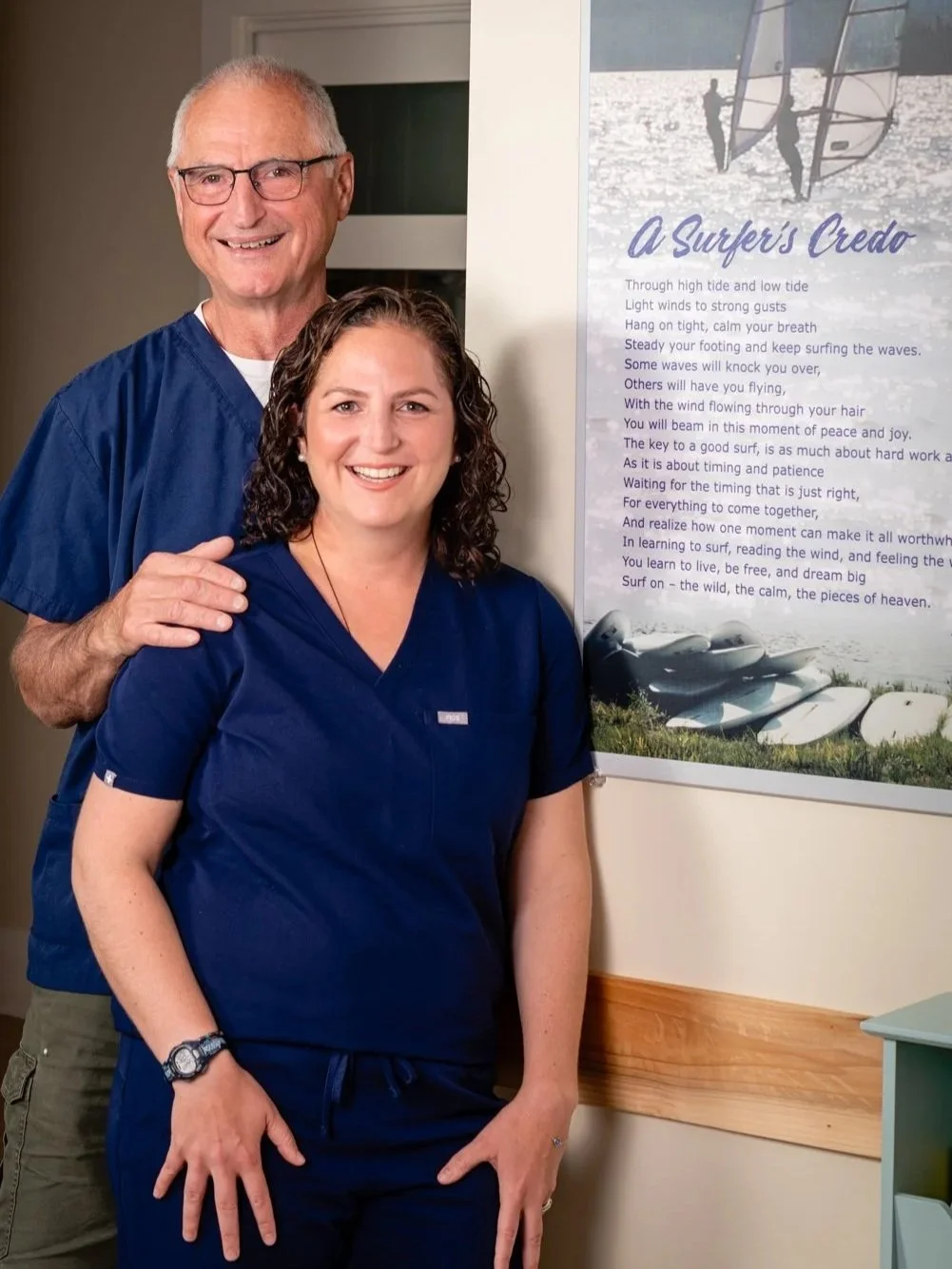 Two healthcare workers, a man and a woman, standing in front of a wall with a surfing-themed poster that has a poem titled 'A Surfer's Creed.' The man, with short gray hair and glasses, is dressed in blue scrubs and has his hand on the woman's shoulder. The woman, with curly brown hair, is also dressed in blue scrubs and is smiling at the camera.
