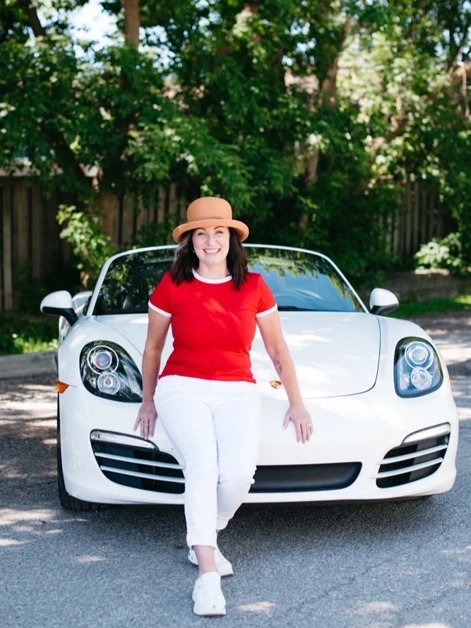 Woman in red shirt and white pants sitting on the hood of a white sports car outside, with green trees and a wooden fence in the background.