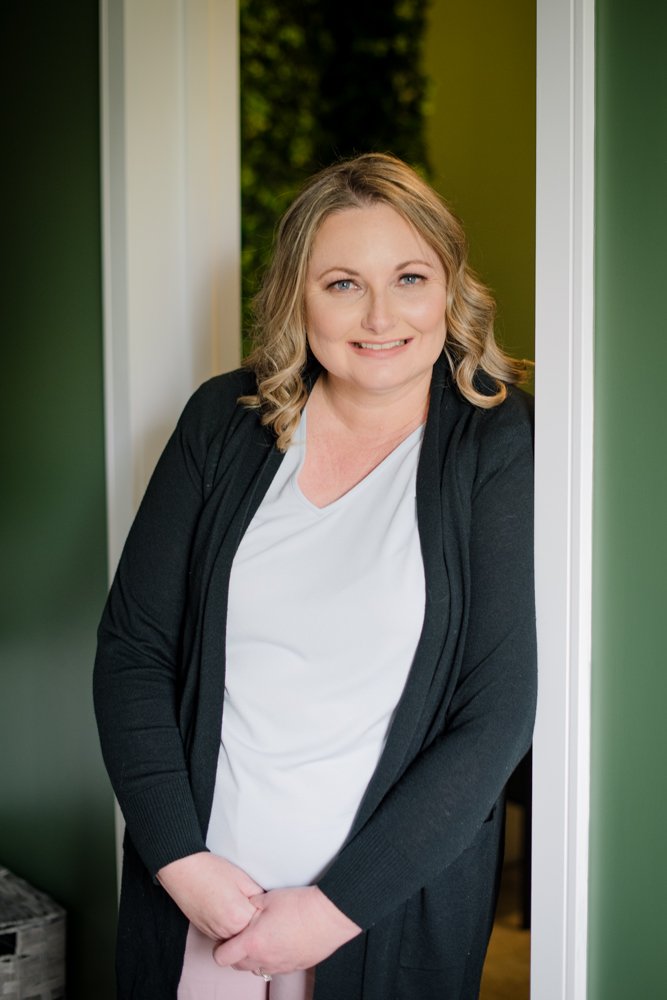 A woman with blonde hair wearing a white shirt and black cardigan standing in a doorway, smiling at the camera.