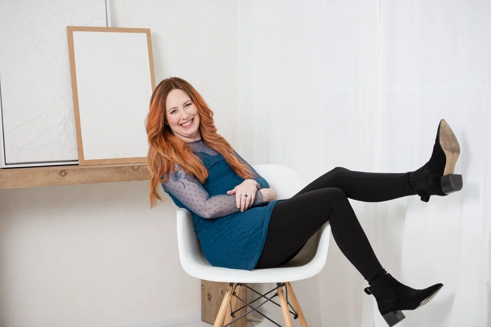 A woman with red hair, wearing a blue dress and black leggings, sitting in a white chair with her legs extended and crossed, smiling and posing in a bright room.