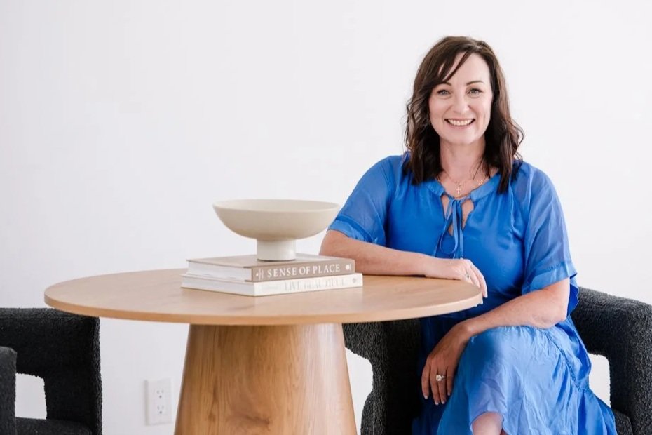 Woman with brown hair in a blue dress sitting on a black chair next to a round wooden table with books and a decorative bowl on top.