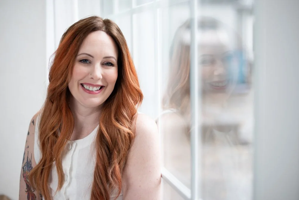 A woman with long, red hair smiling near a window, with her reflection visible in the glass.