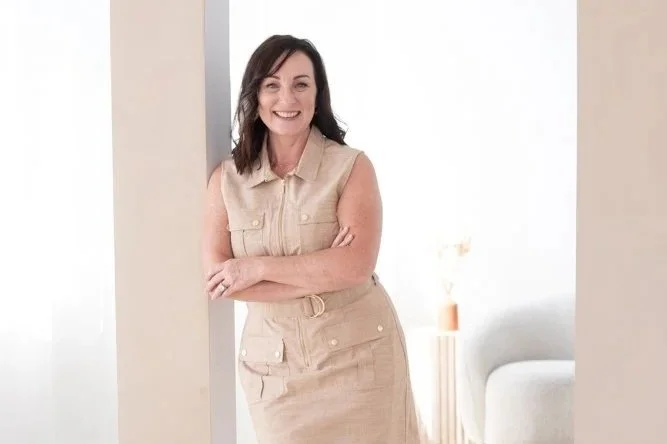 A woman with shoulder-length dark hair, smiling, wearing a sleeveless beige dress with a belted waist, standing in a bright indoor space.