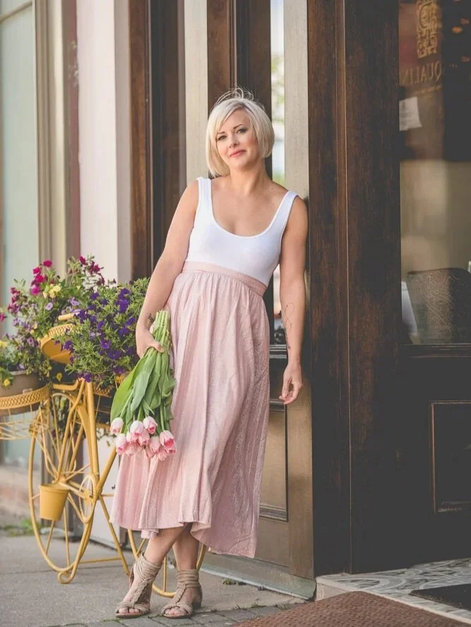 Woman with short blonde hair in a white tank top and pink pleated skirt holding pink tulips stands outside a shop with a flower cart nearby.