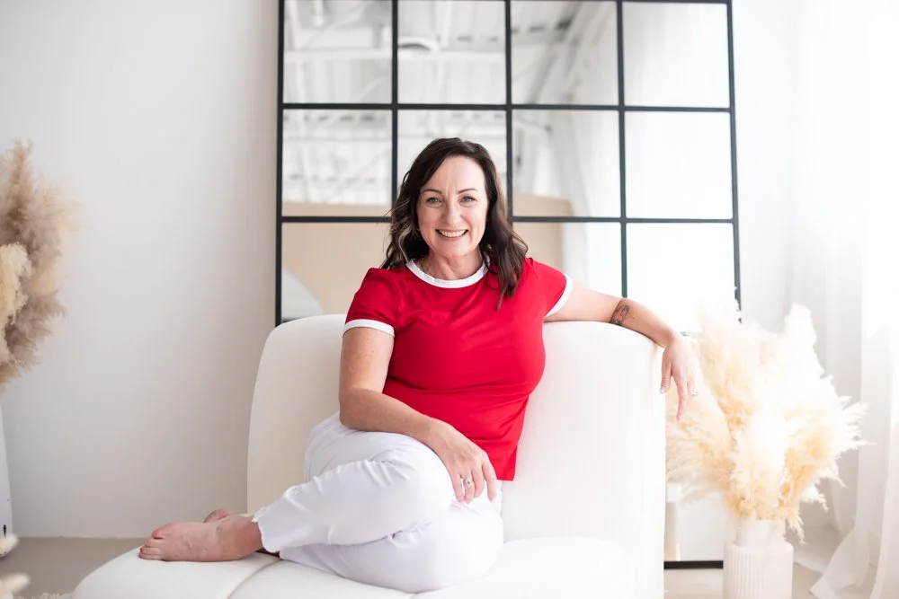 Smiling woman with dark hair wearing a red shirt and white pants sitting on a white sofa in a bright, modern room with decorative pampas grass in a vase.