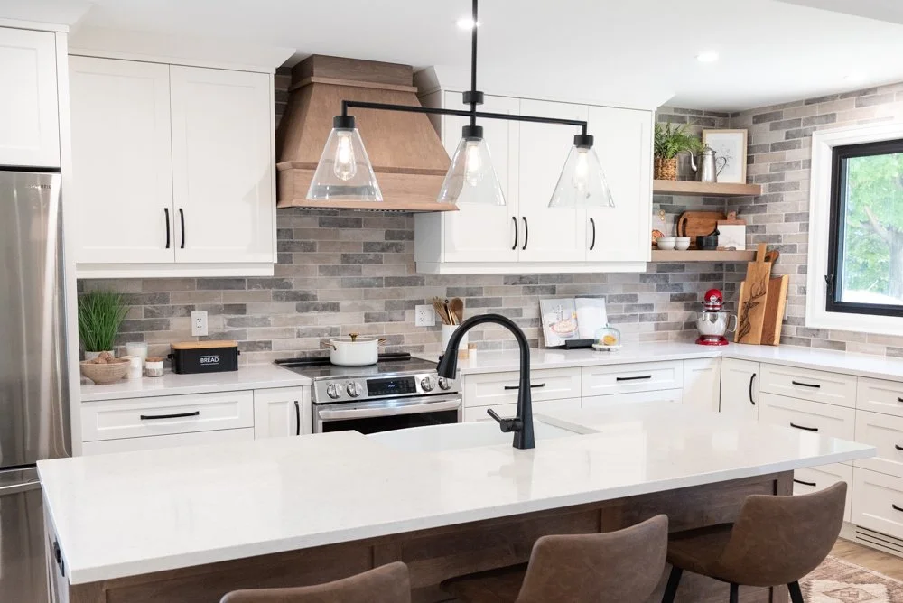 Modern kitchen with white cabinets, a large island with a white countertop, and a black faucet. Stainless steel refrigerator, gray brick backsplash, and open wooden shelves with plants and kitchenware. Pendant lighting over the island, window showing green trees outside.