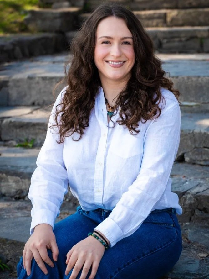 A young woman with long curly hair sitting on stone steps outdoors, smiling, wearing a white shirt and blue jeans.