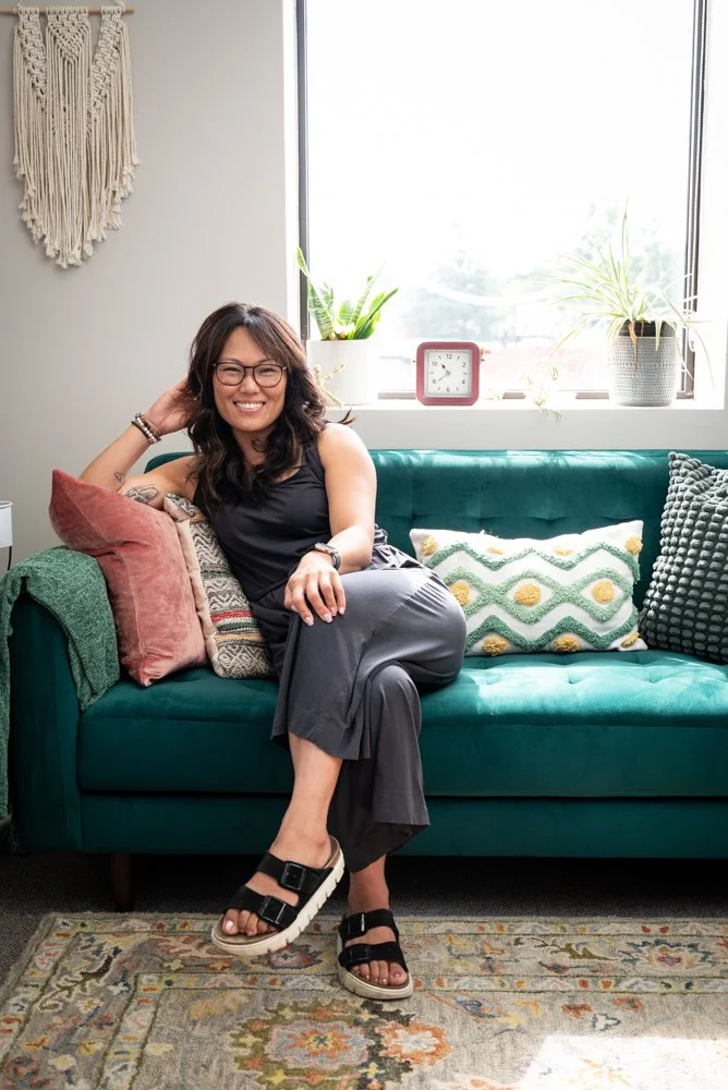 Woman with dark hair, glasses, black top, and gray pants sitting on a teal sofa, smiling. The sofa has decorative pillows, with a pink pillow on her left. Behind her, a window with sunlight, potted plants, a clock, and a wall hanging decorate the room. A patterned rug covers the floor.