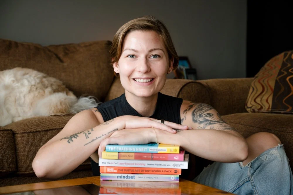 A woman with short hair, tattoos, and a black sleeveless top smiling while resting her arms on a stack of books on a wooden table. A dog is lying on a couch in the background.