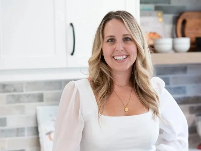 A woman with blonde hair smiling in a kitchen with white cabinets and a tiled backsplash.