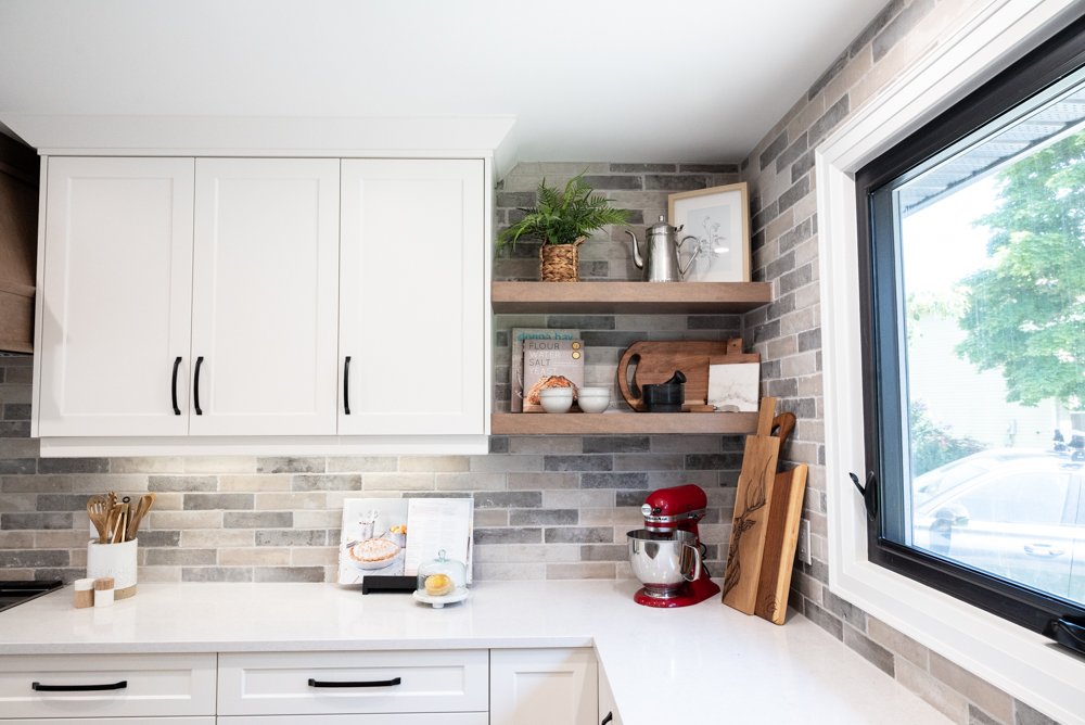 Kitchen countertop with a red stand mixer, wooden cutting boards, and a small picture frame. Open shelves with a plant, a watering can, bowls, a book, and kitchen utensils. A window showing trees outside.