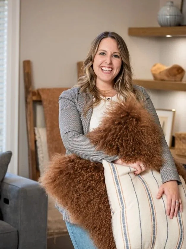 A woman with long wavy blonde hair smiling and holding a fluffy brown dog, sitting on a striped pillow.