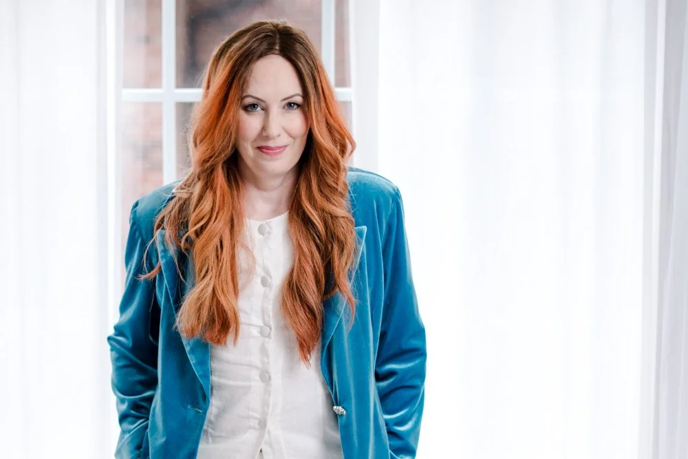 A woman with long red hair and fair skin standing indoors in front of windows with white curtains, wearing a blue blazer and a white top.