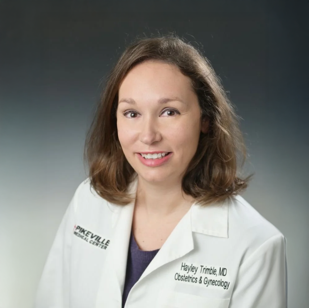 A woman with shoulder-length brown hair and light skin smiling in a white medical coat with name and credentials, standing against a gradient dark background.