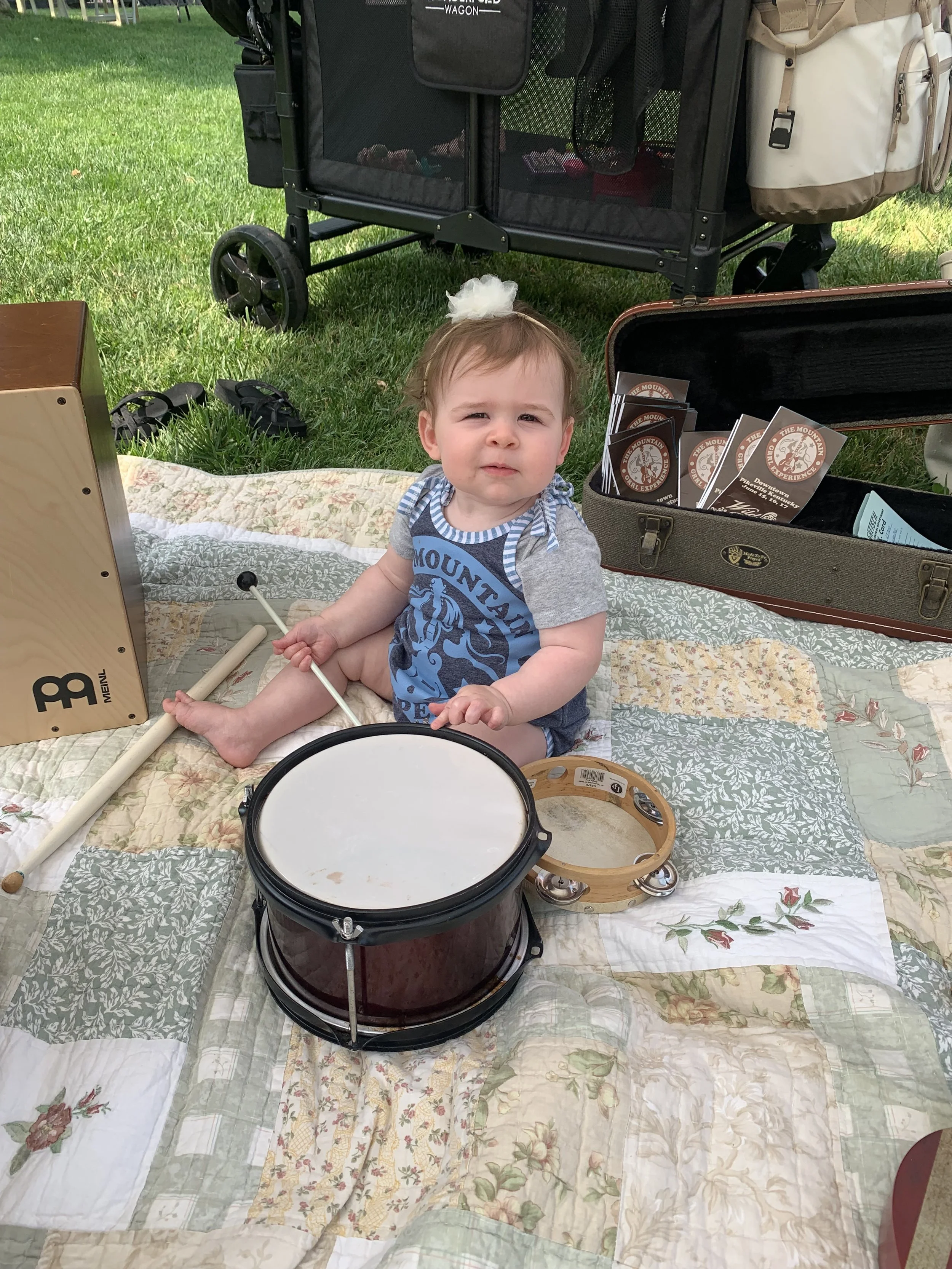 A young child sitting on a quilted blanket outdoors, surrounded by musical instruments including a drum, tambourine, and a small wooden percussion instrument. There is a stroller and some pamphlets in the background.
