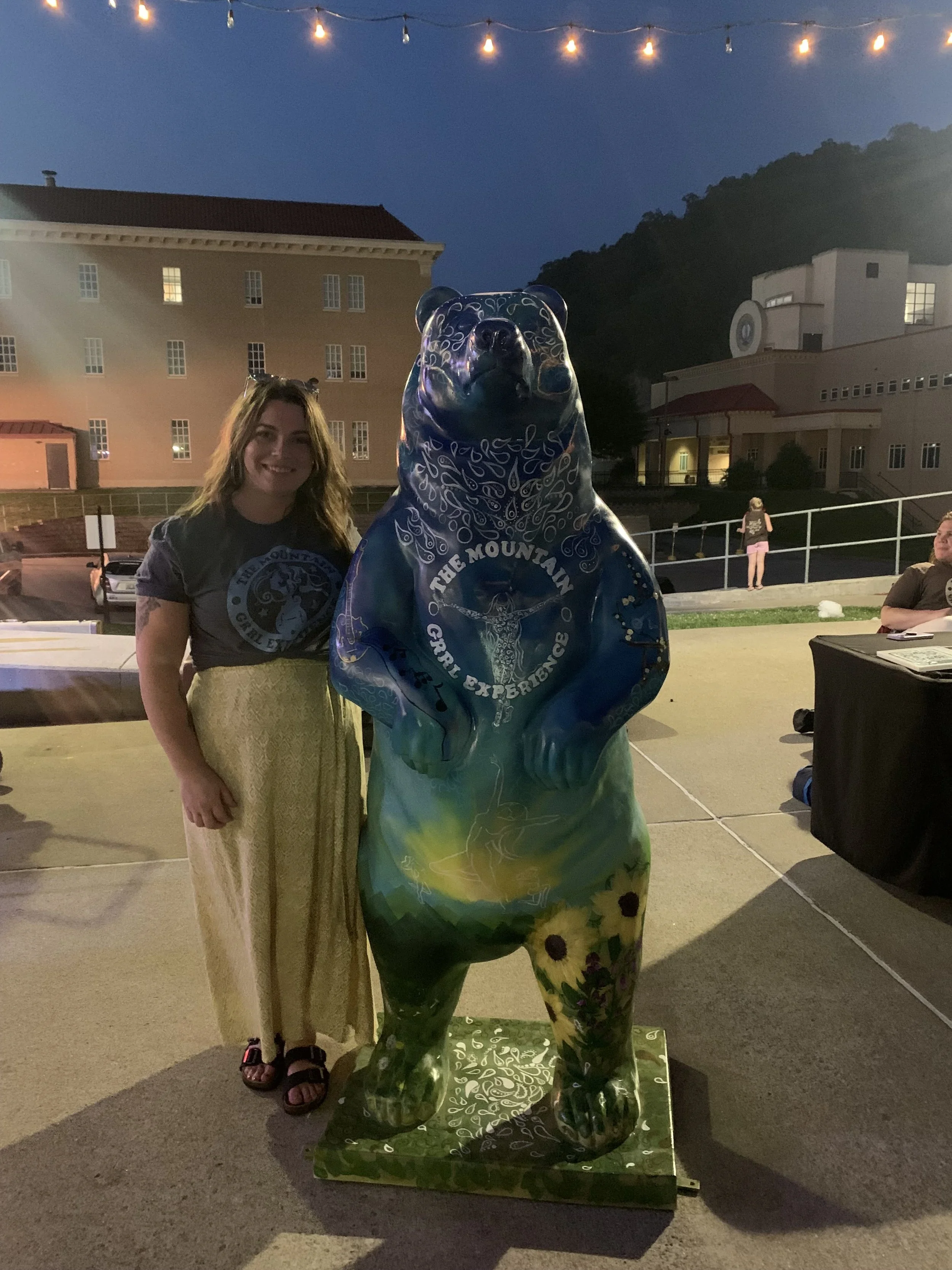 A woman standing next to a painted bear sculpture outdoors at dusk, with string lights overhead and a building and hills in the background.