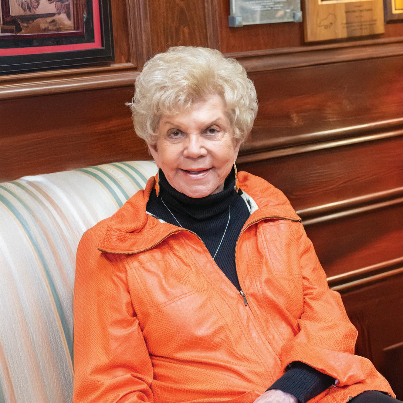 An elderly woman with short curly blonde hair, wearing an orange jacket and black turtleneck, sitting on a striped beige and light blue cushioned bench in a wood-paneled room.