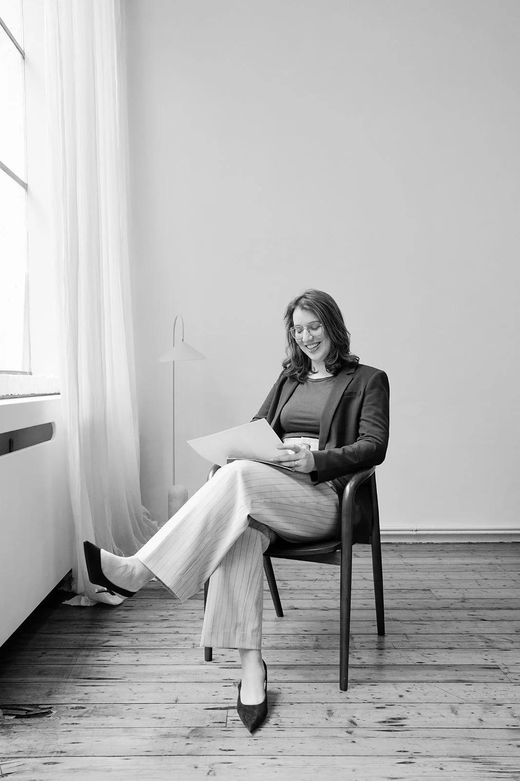 Charlotte zit op een stoel bij het raam, leest documenten en glimlacht; ze draagt een blazer en een gestreepte broek in een minimalistische kamer met houten vloeren.