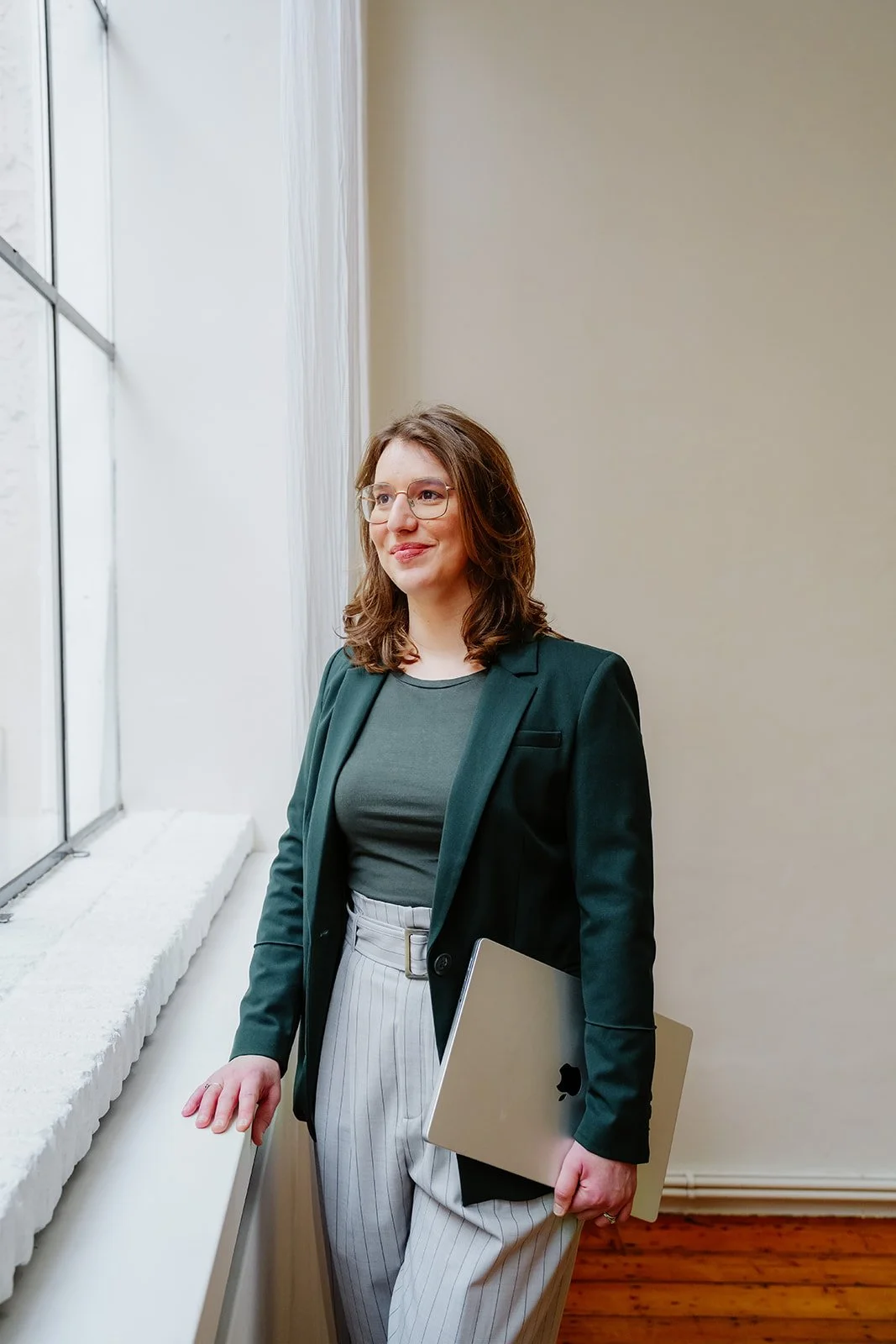 Charlotte, gekleed in een donkergroene blazer en een licht gestreepte broek, met haar laptop in haar handen, staande bij een raam waar natuurlijk licht naar binnen valt.