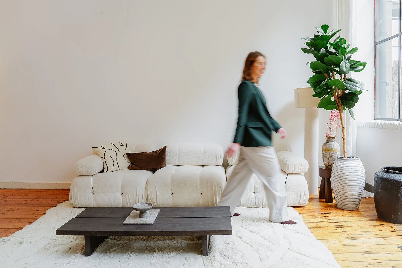 Charlotte loopt langs een crèmekleurige bank in een lichte woonkamer met houten vloeren, een salontafel, een grote kamerplant en een raam met witte gordijnen.