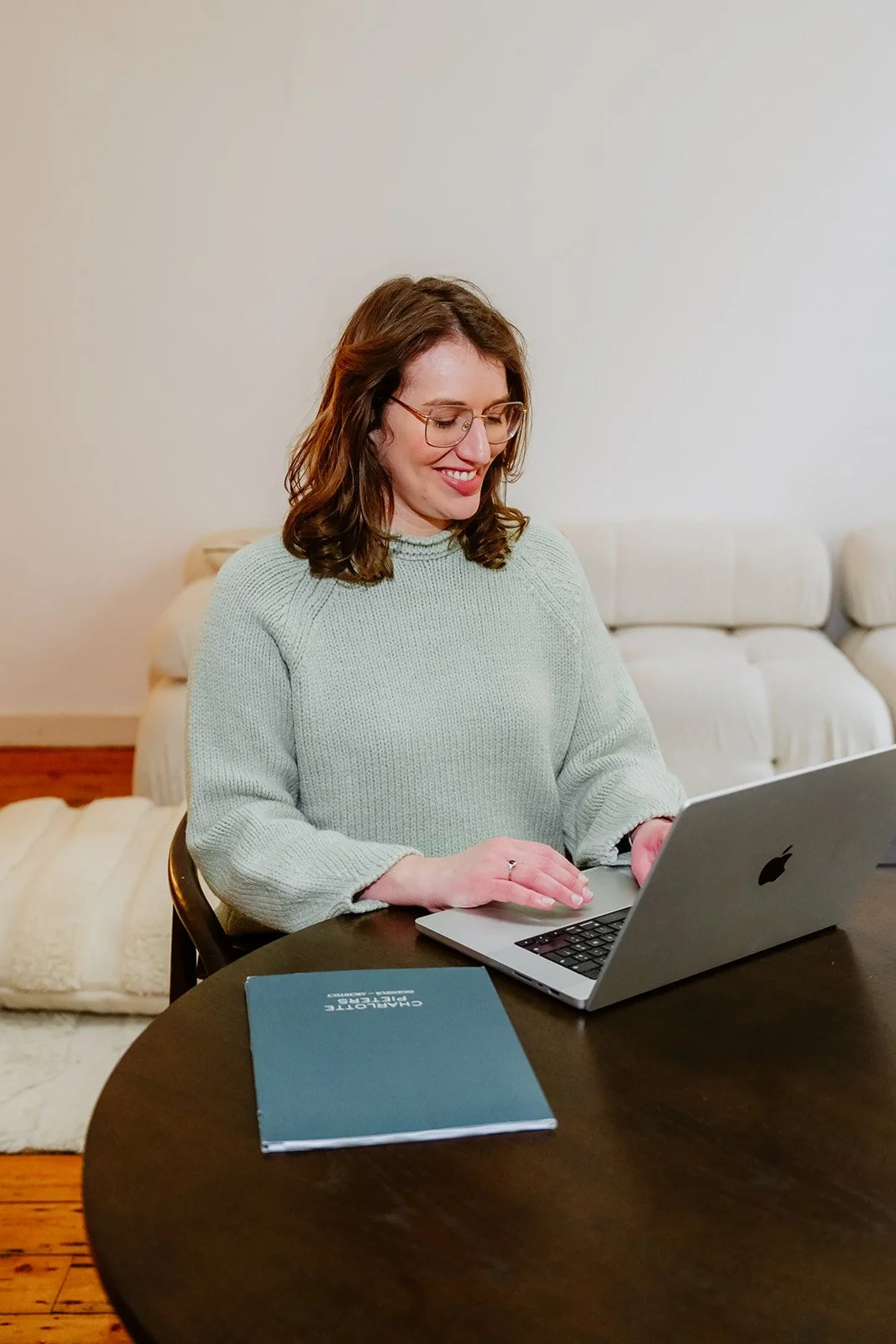 Charlotte, gekleed in een lichtgroene trui, zit aan een donkere houten tafel. Ze glimlacht en werkt op haar laptop, met een blauw notitieboekje voor zich, in een kamer met een beige bank en een crèmekleurige deken.