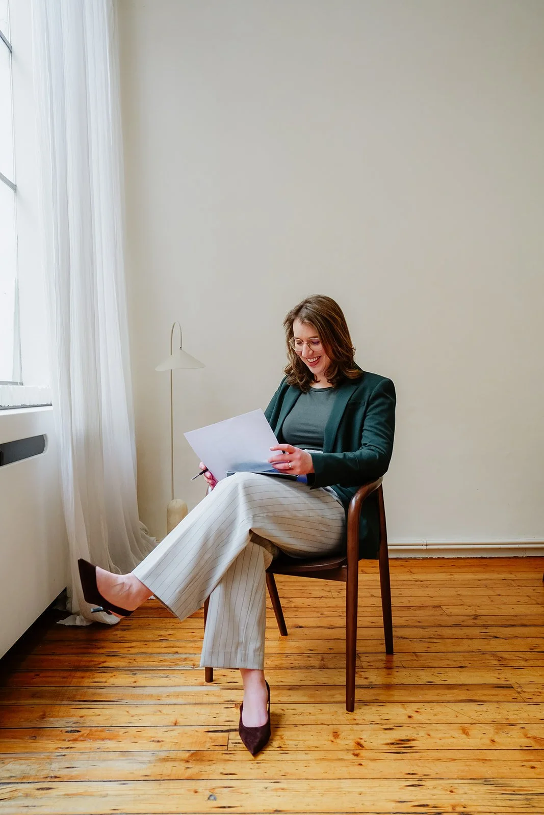 Charlotte zit op een houten stoel bij het raam; ze glimlacht en leest een document met een pen in de hand, in een kamer met witte muren en een houten vloer.