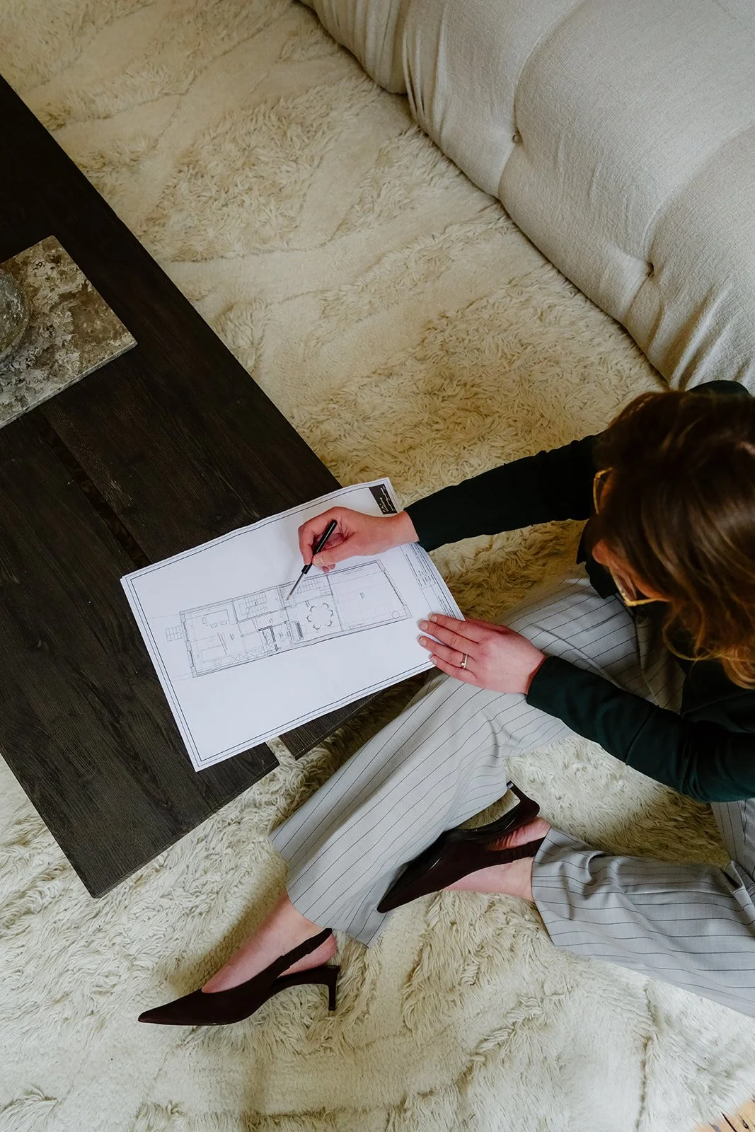 Charlotte zit op een crèmekleurig, hoogpolig tapijt en bekijkt bouwtekeningen aan een donkere houten tafel in een crèmekleurige woonkamer.