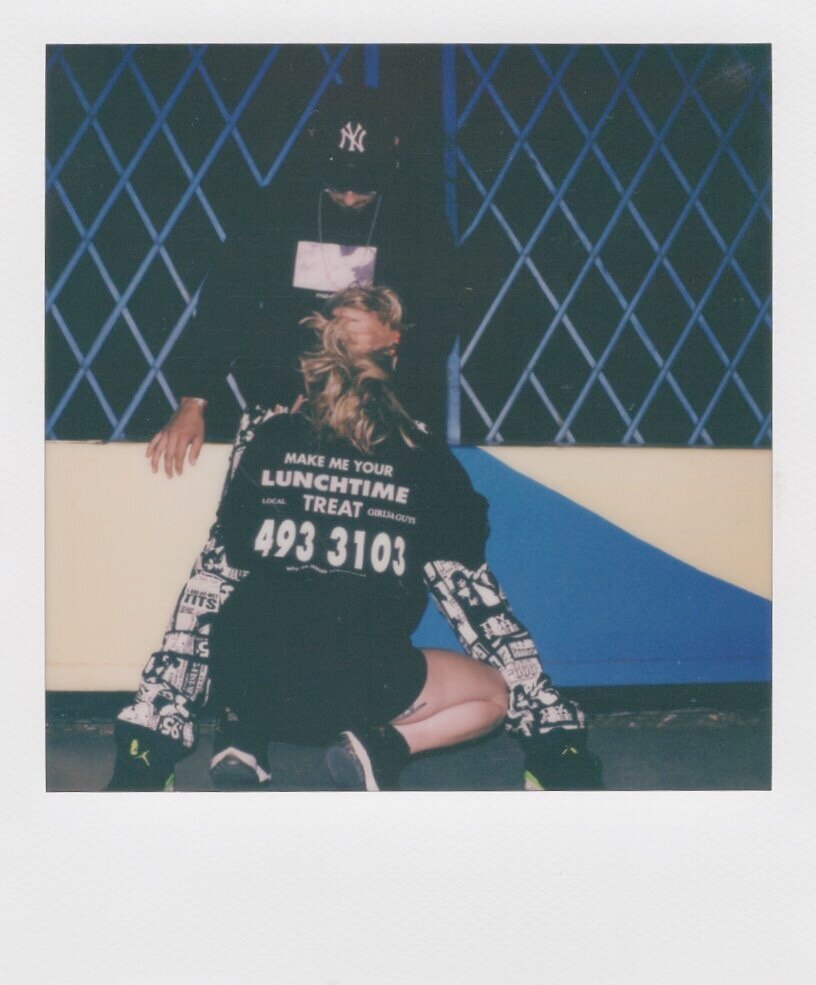 A person kneeling on the ground with their head down, wearing a black and white patterned jacket and black shorts, with a blanket over their lap. Behind them, a man wearing a black New York Yankees cap and hoodie stands with one hand on the wall. The