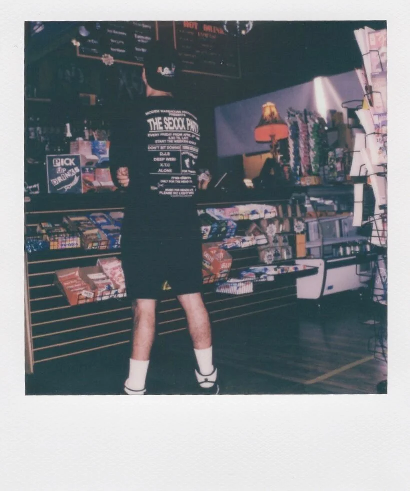 Person standing in a convenience store, wearing a black band T-shirt, black shorts, white socks, and sandals, with shelves of snacks, drinks, and other items behind him.