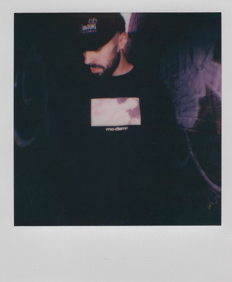 A man with a beard wearing a black baseball cap and black shirt, looking down, with a photo on his shirt and a dark background.