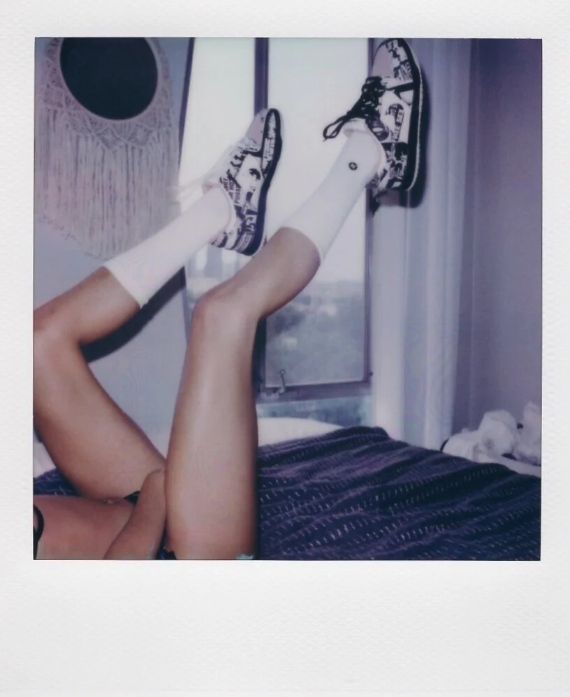Person lying on bed with legs raised and resting on a window, wearing sneakers and socks, in a bedroom.