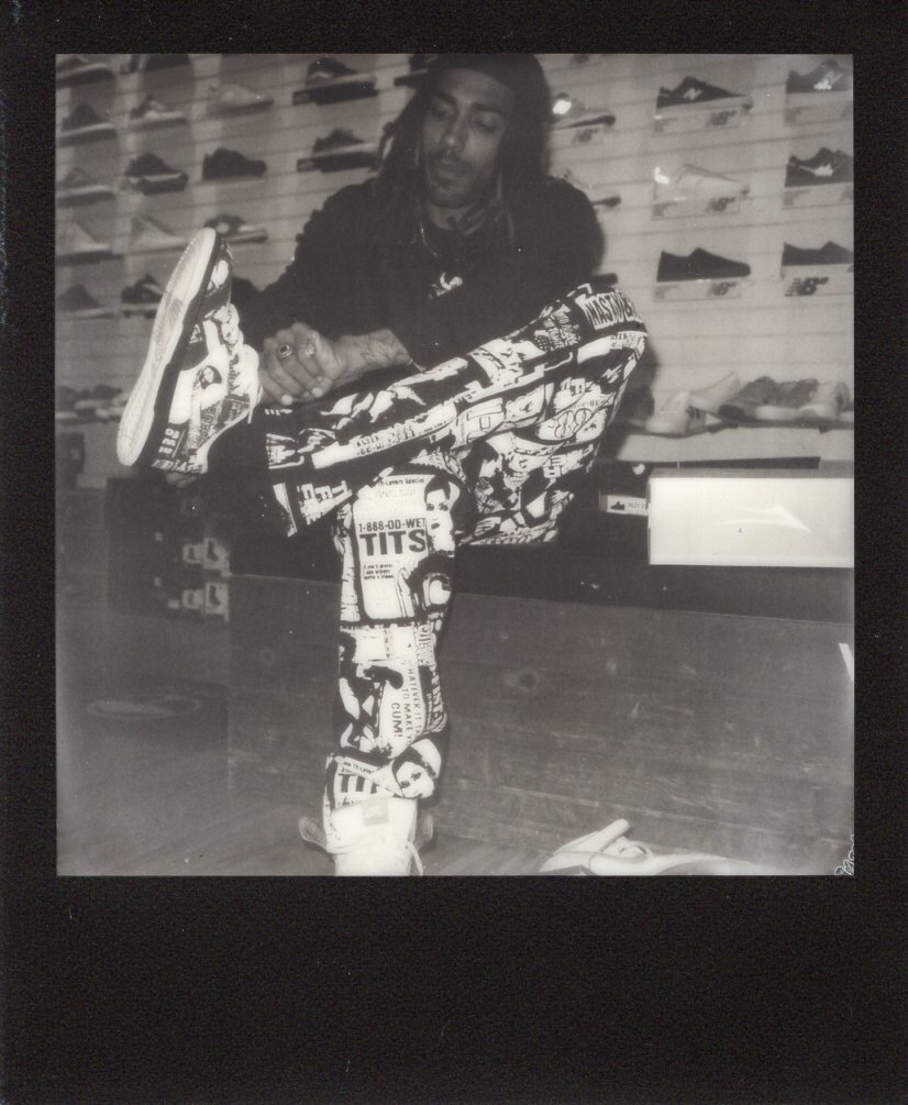 A man with dreadlocks in a shoe store holding his leg up to show off a colorful comic-style painted sneaker.