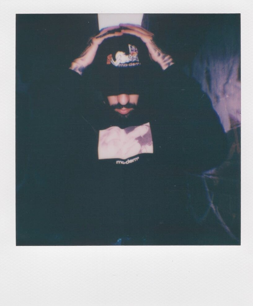 Person wearing a black hoodie with the word 'mo-dern' and a graphic, holding hands on their head with hands visible at the top of the image.