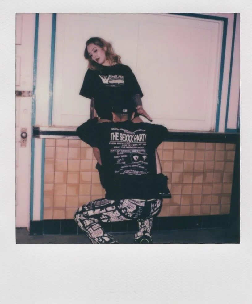 A woman with wavy hair and makeup standing behind a man sitting on a counter. The woman is looking at the man who has his back to the camera. The woman is wearing a black T-shirt, and the man is wearing a black T-shirt with a graphic and black-and-wh