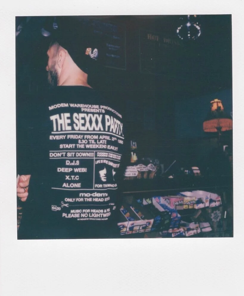 A person wearing a black t-shirt with white text and a graphic on the back, standing in a dimly lit bar or club setting with arcade games and a decorative lamp in the background.