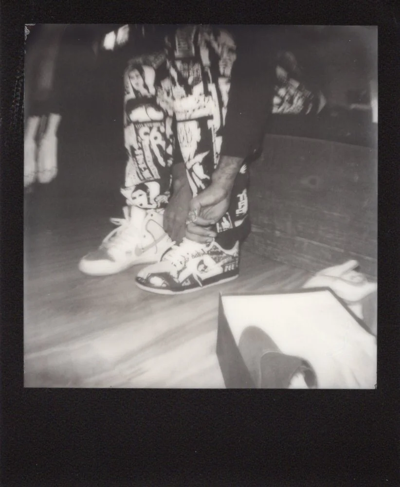 A person is tying the shoelaces of their sneaker, which has a colorful, patterned design, while standing on a wooden floor. The person is wearing patterned pants.