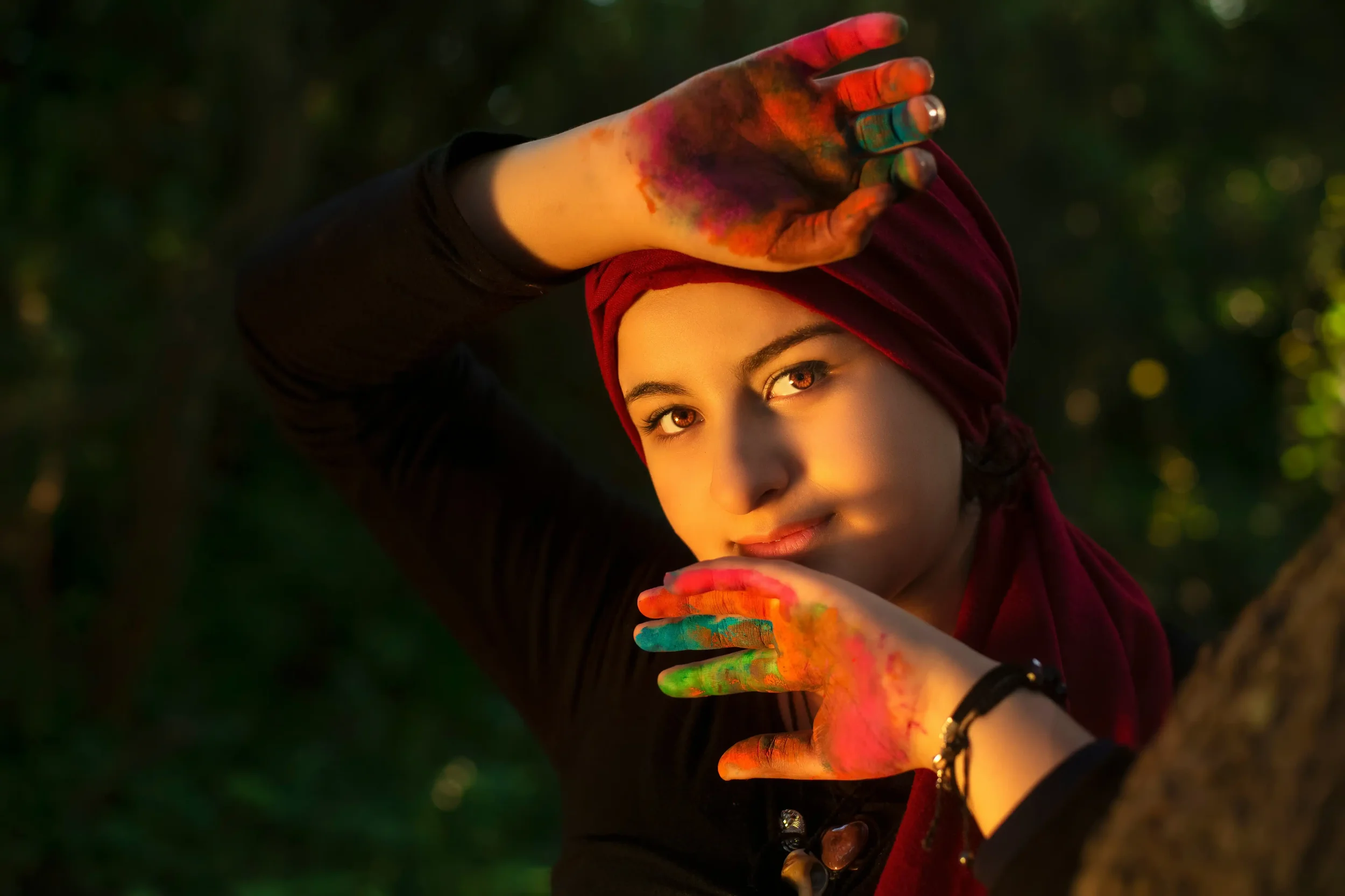 A person wearing a red headscarf poses outdoors with colorful paint on their hands, holding them near their face. Sunlight highlights their features, and the background is blurred greenery.