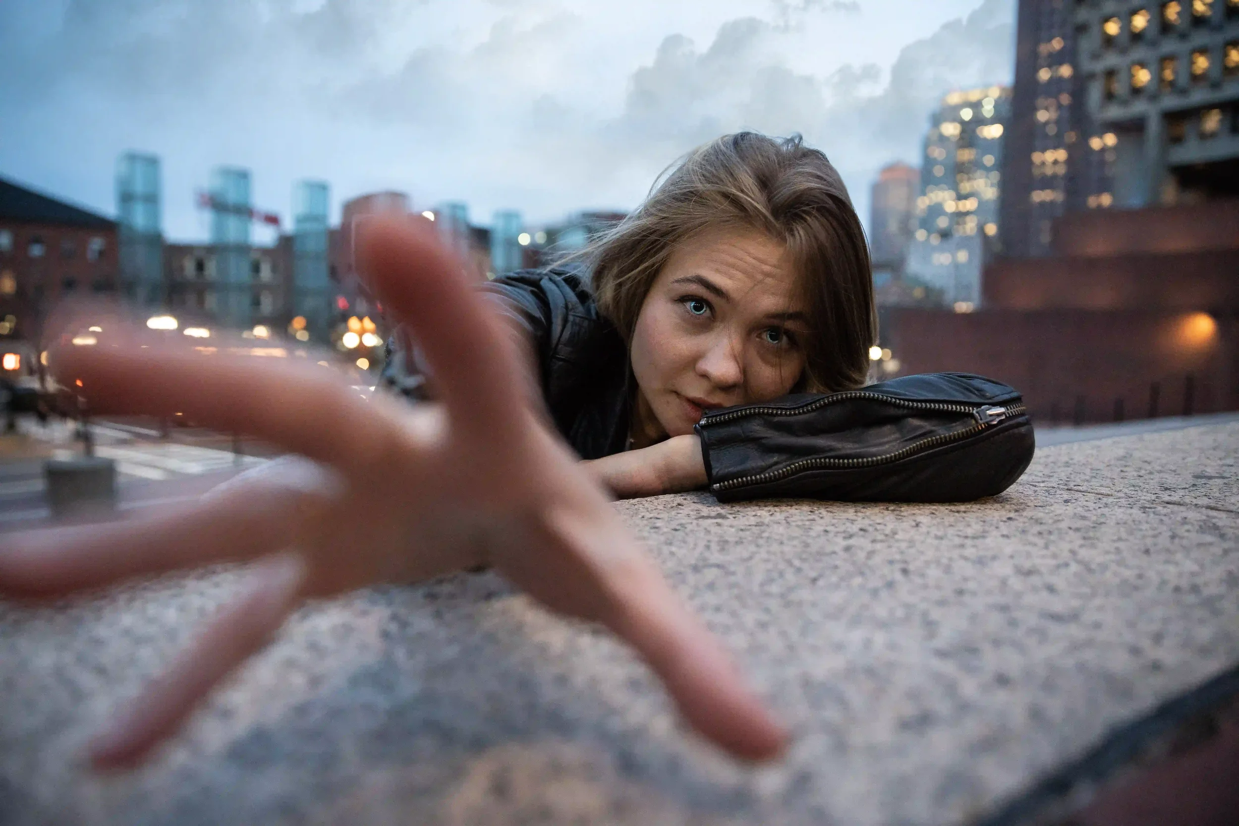 A woman in a black jacket leans on a stone surface, reaching toward the camera with one hand. City buildings and lights are blurred in the background at dusk.