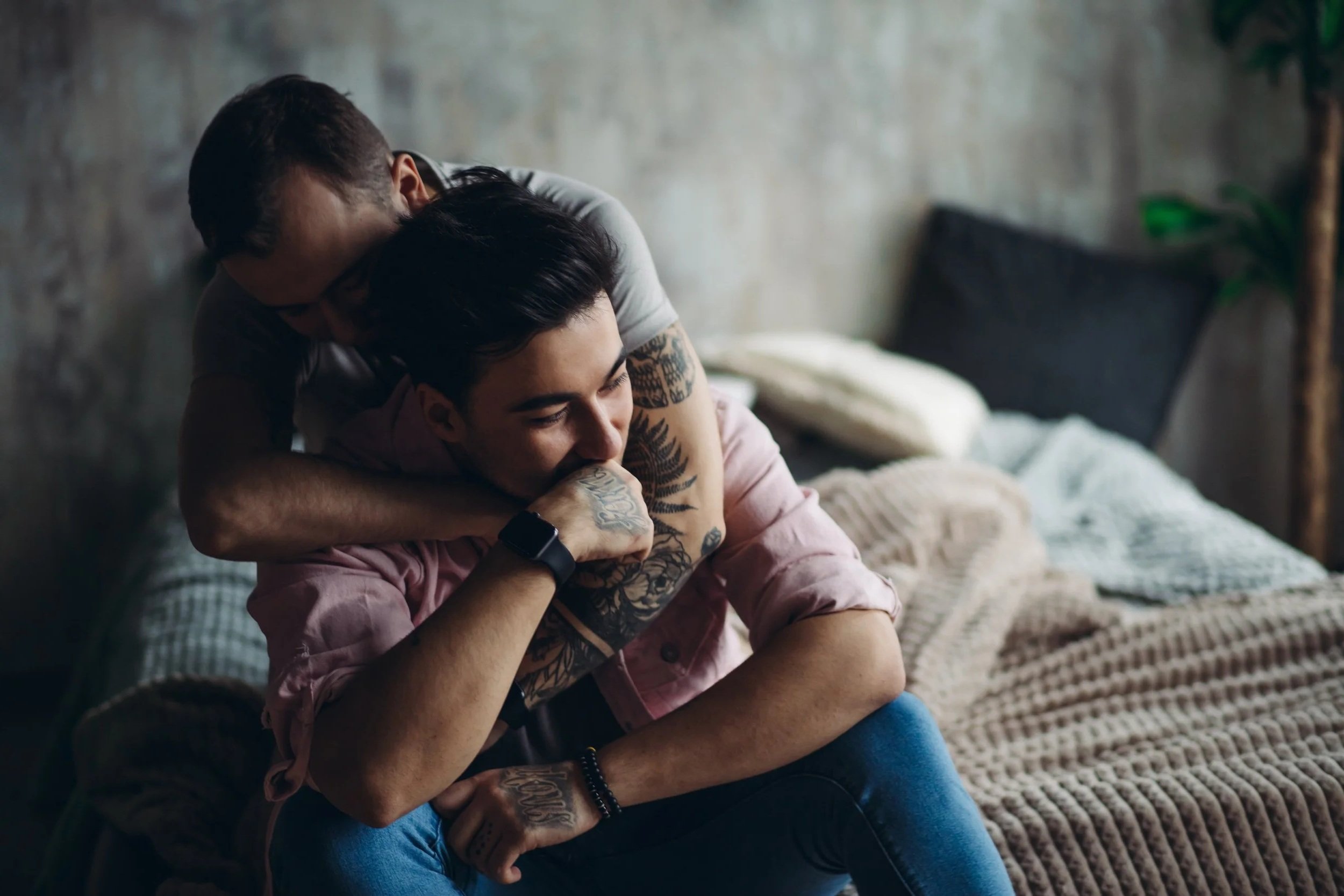 A man sits on the edge of a bed while another man embraces him from behind.
