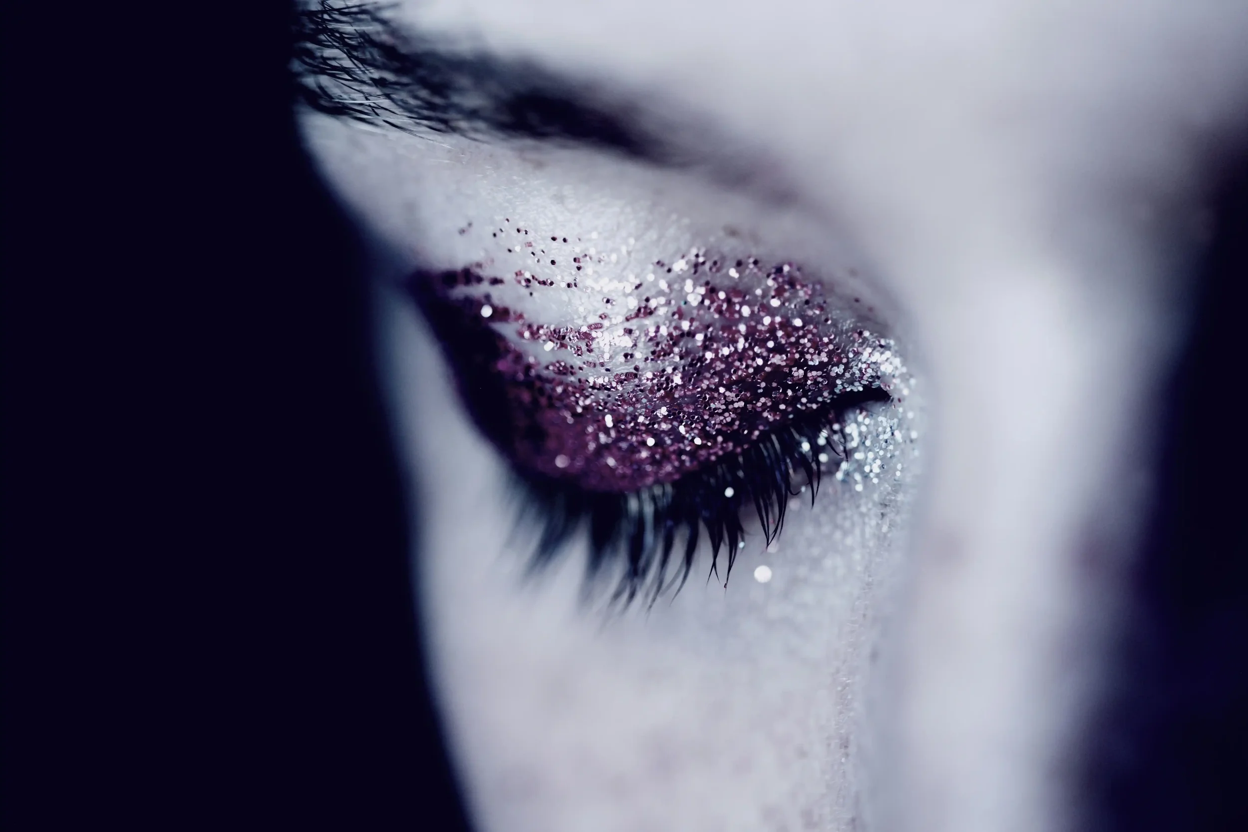 A close-up of a closed eye with long eyelashes, featuring shimmering pink and silver glitter eyeshadow on the eyelid.