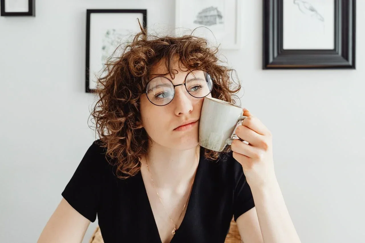 A woman with curly hair and round glasses holds a mug against their cheek while looking away.