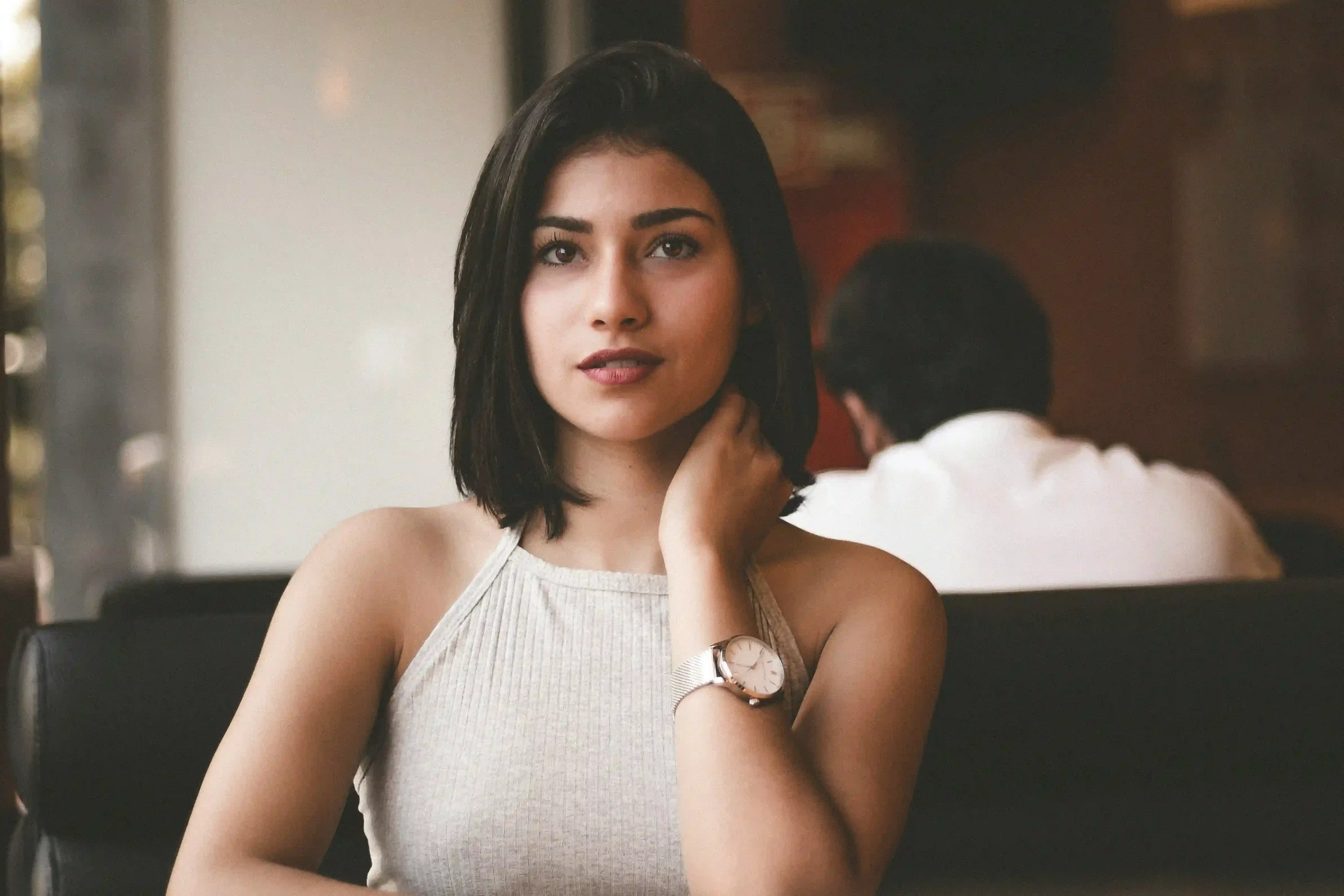 A woman with medium-length dark hair sits at a table indoors, wearing a sleeveless, light gray top and a watch on her left wrist. She looks at the camera with a neutral expression. A person sits in the background, facing away.