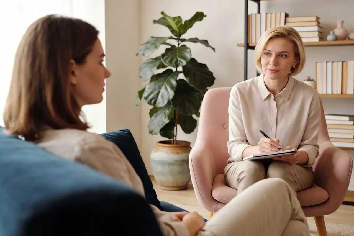 A woman sits on a couch talking to another woman who is taking notes on a clipboard; they are in a cozy room with a plant and bookshelves, suggesting a therapy or counseling session.