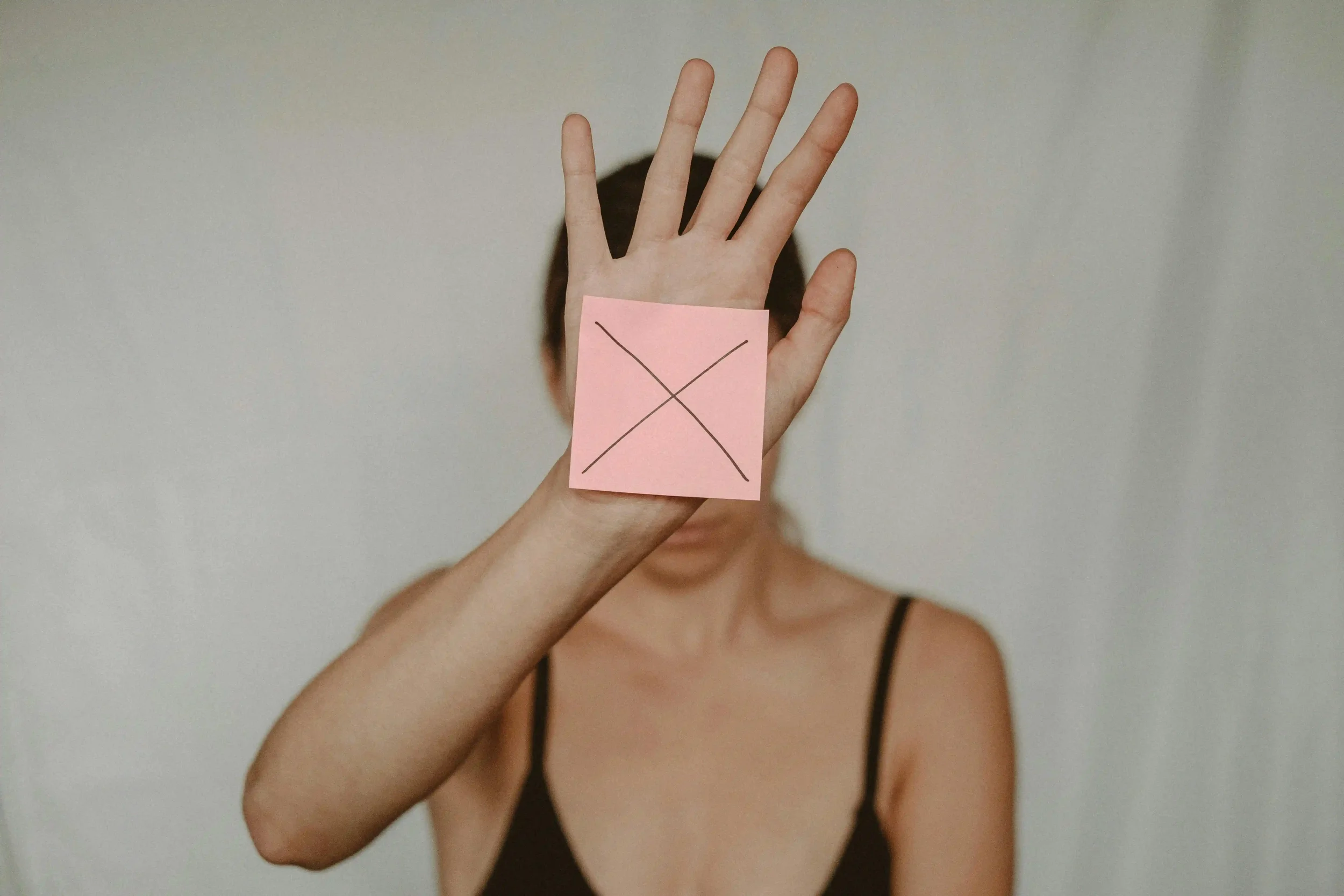 A person holds up their hand with a pink sticky note displaying a large black X, covering their face. The background is neutral, and only the upper body is visible.