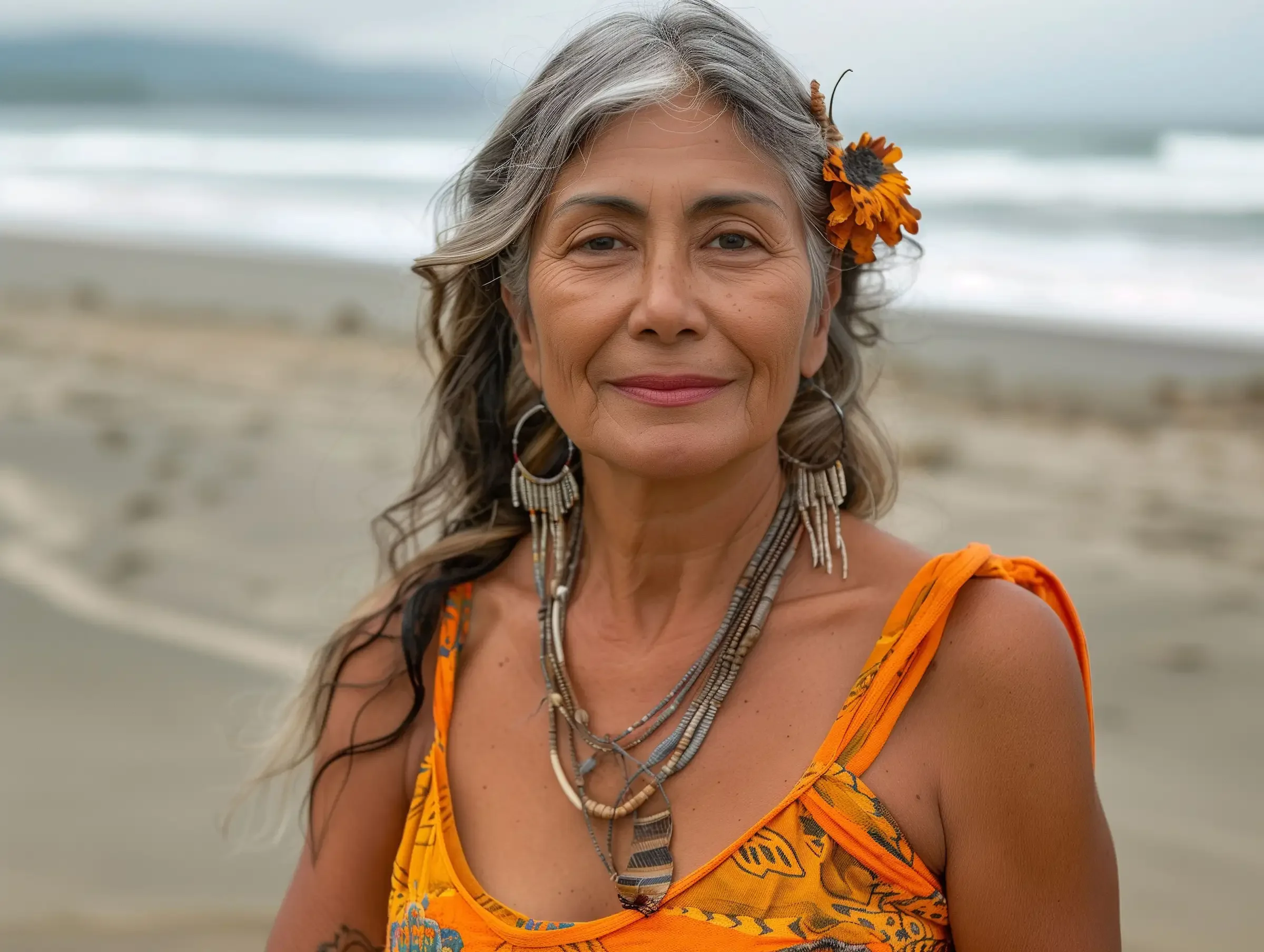 A smiling older woman with long gray hair and a flower tucked behind her ear stands on a beach, wearing a bright orange patterned dress and layered necklaces. The ocean and cloudy sky are in the background.