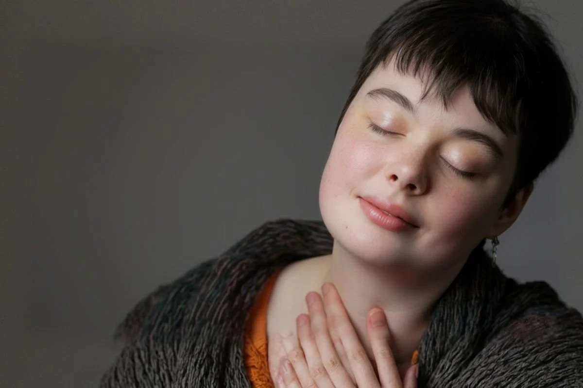Close up portrait of a person with short dark hair resting a hand on their throat and tilting their face upward with their eyes closed. Soft lighting and a calm expression support content about how to calm your nervous system.