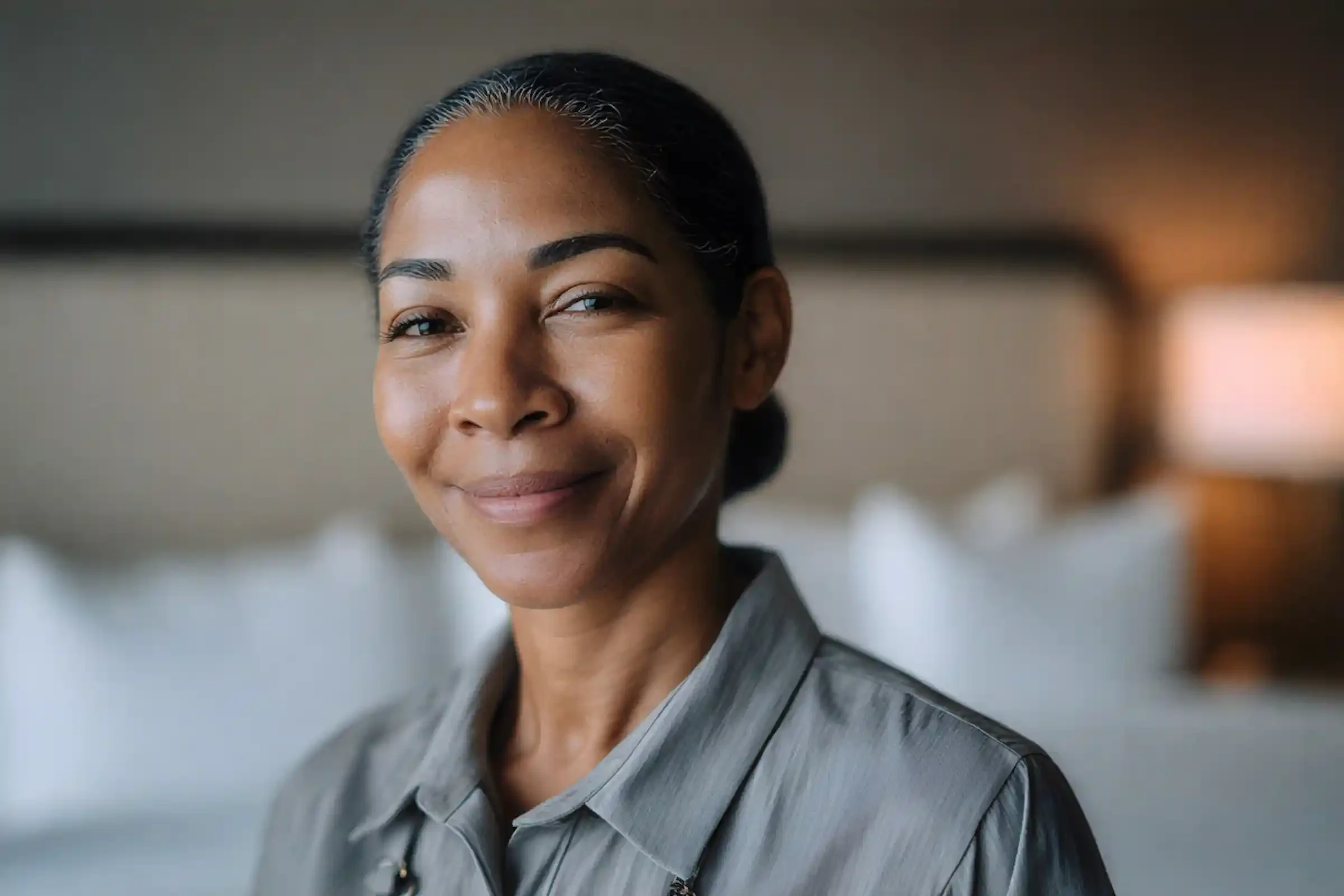 A woman with dark hair pulled back, wearing a gray collared shirt, smiles softly while standing indoors in a softly lit room with blurred white pillows and a lamp in the background.